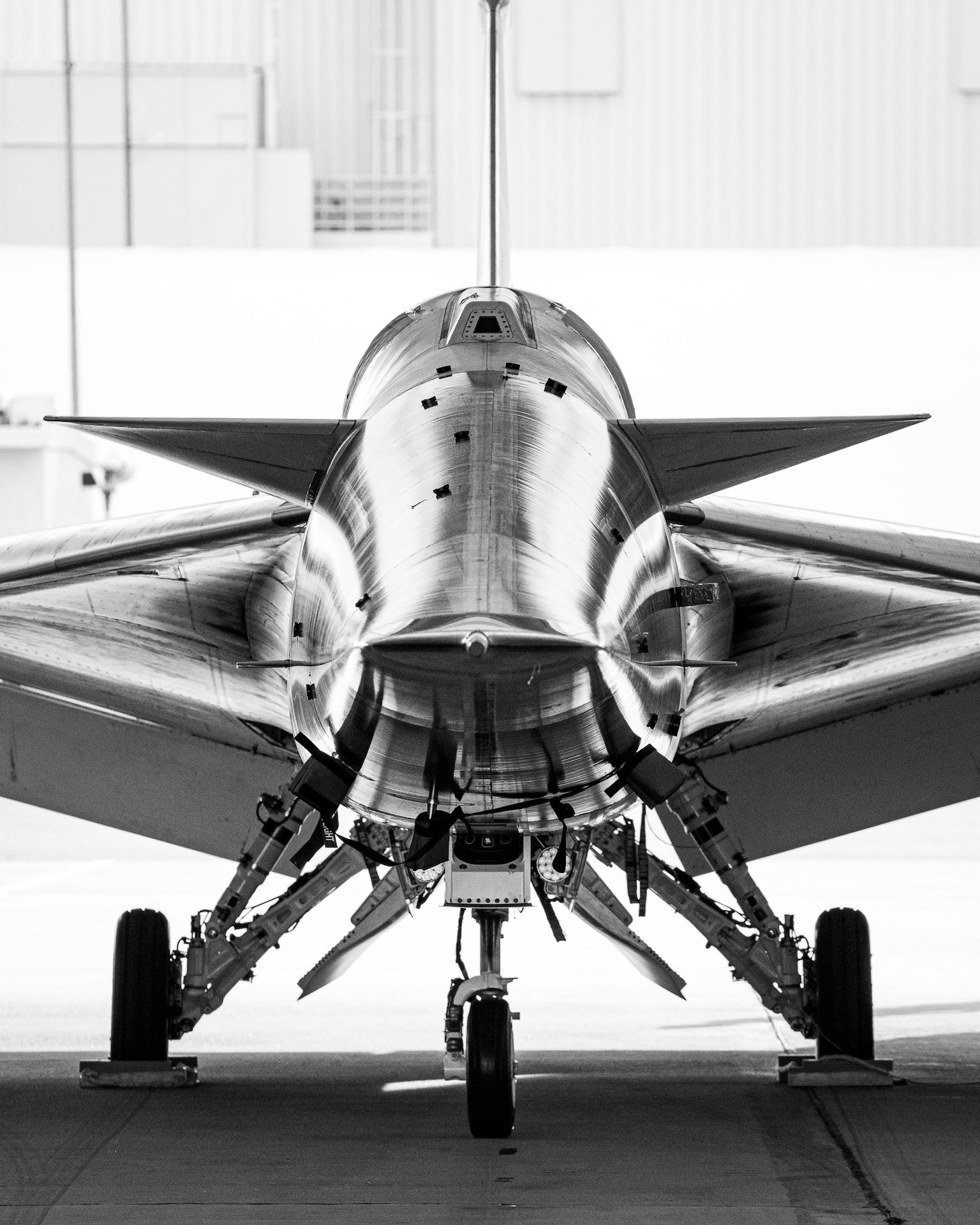 X-59 süpersonik jetin kafa kafaya siyah beyaz görüntüsü. Jet, üç tekerlekli iniş takımları üzerinde oturuyor ve jetin gövdesi ile kanatlarının keskin, şık açıları, aracın kıvrımlarını ve ayrıntılarını vurgulayarak düzgün bir ışık olarak yansıyor.