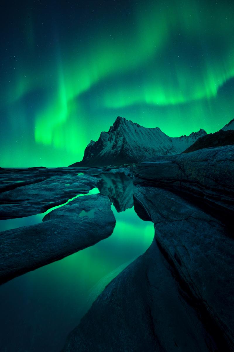 Aurora borealis'in yeşil dalgaları, yukarıdaki yeşil auroraları yansıtan akan derelerle ön plandaki vadiye kök salmış sivri, kayalık bir dağın üzerinde gökyüzünü çiziyor.