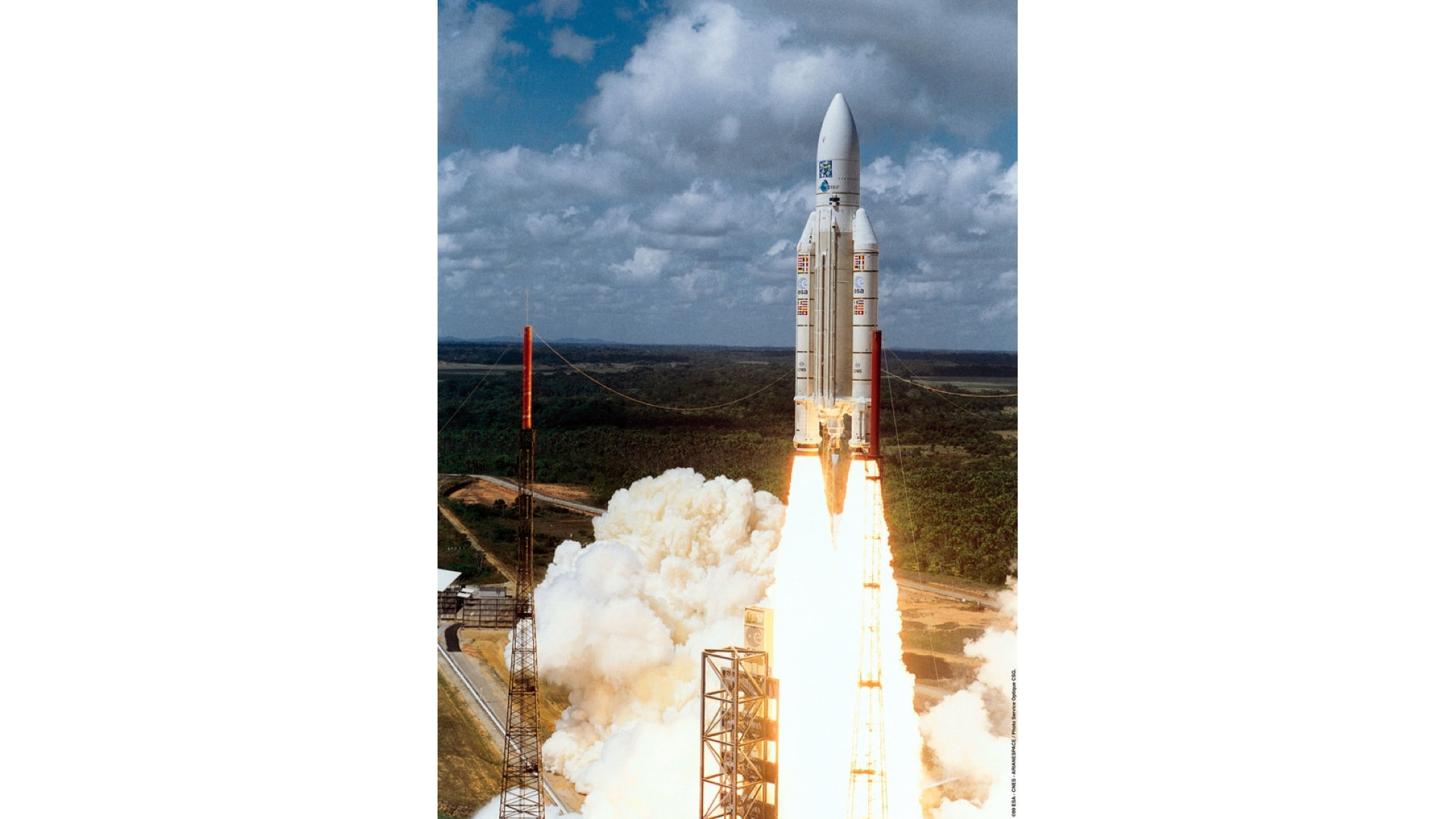 Bir Ariane 5 roketi, yeşil orman ve mavi gökyüzü fonunda fırlatma rampasından havalanıyor.