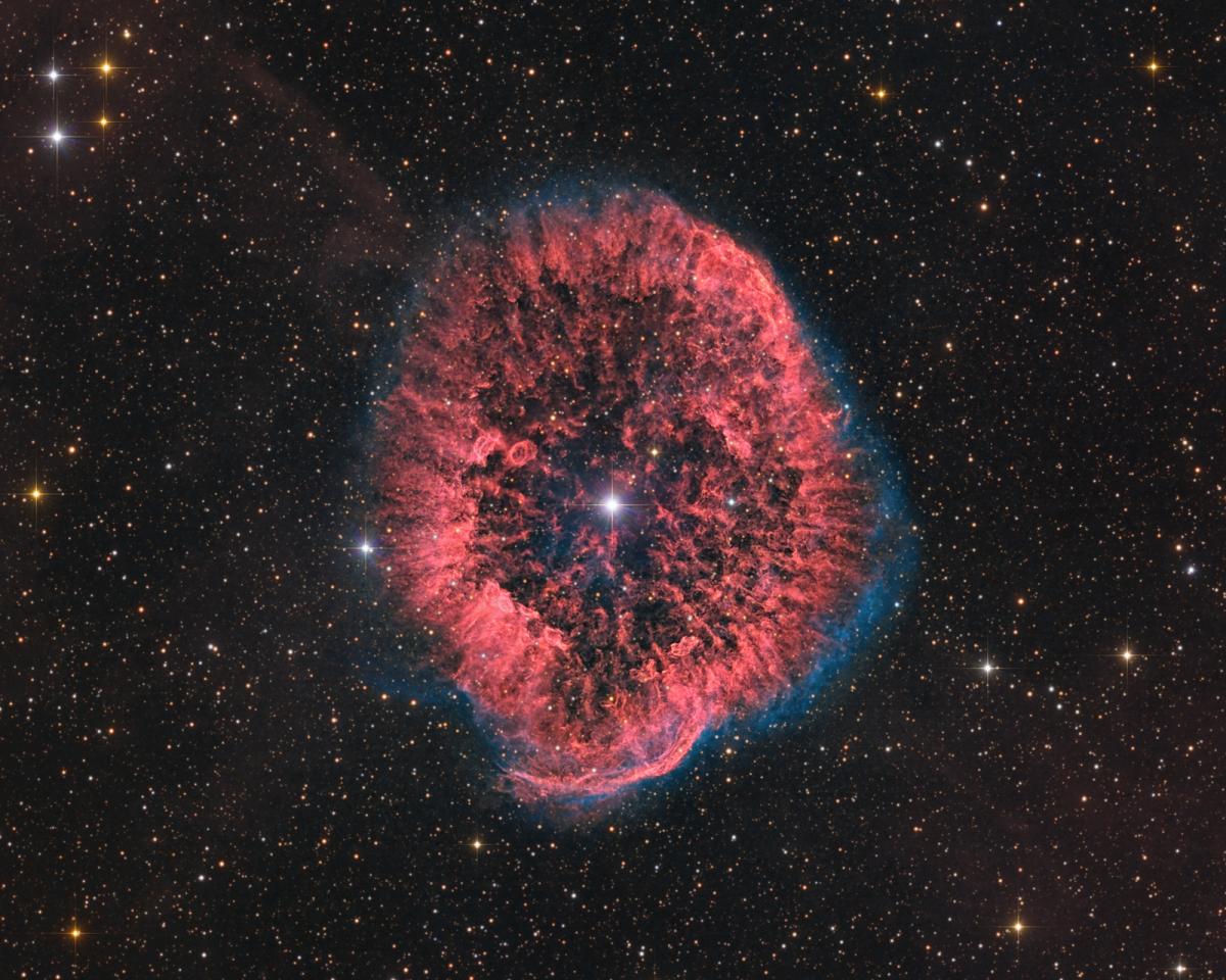 Parlak beyaz bir yıldız, çok katmanlı kırmızı gazlardan oluşan bir nebulanın merkezinde yer alıyor. Uzak yıldızların ve galaksilerin parlak noktaları kırmızı, dairesel patlamayı çevreliyor.